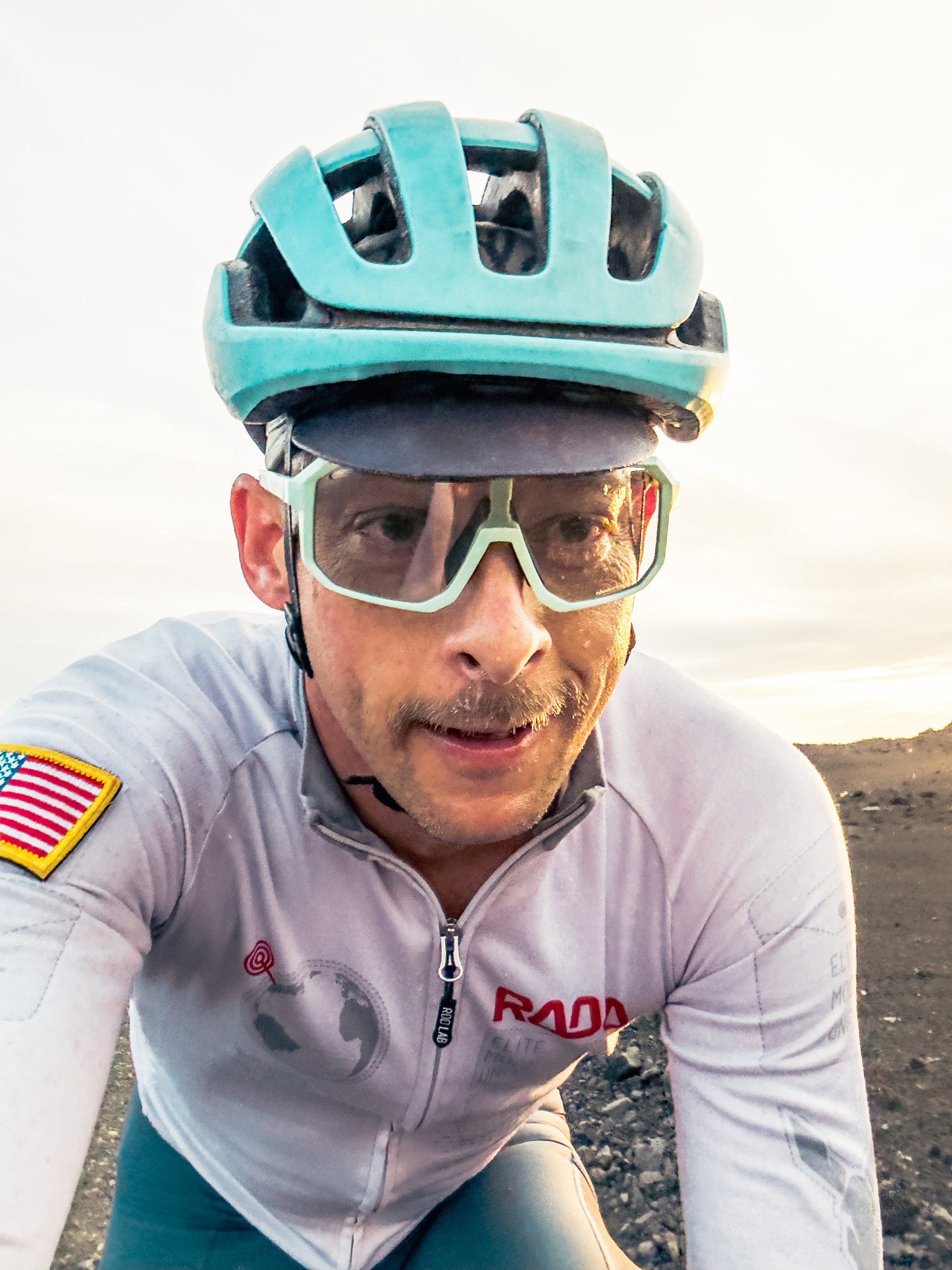 Close-up of a cyclist wearing a teal helmet and glasses, with a serious expression, set in a rocky outdoor environment.