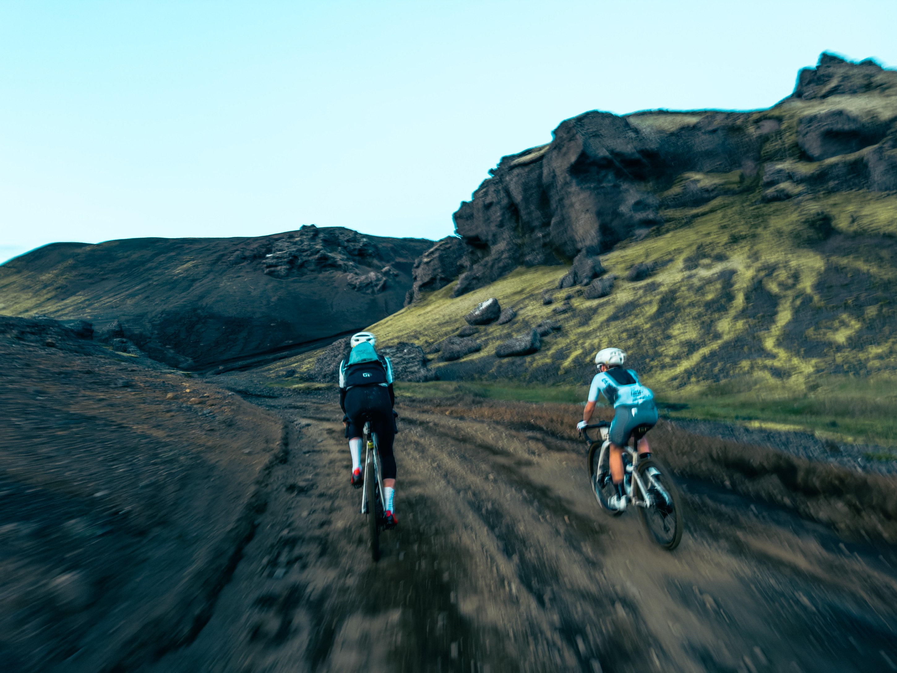 Two mountain bikers riding on a dirt trail surrounded by rugged terrain and hills under a clear blue sky.