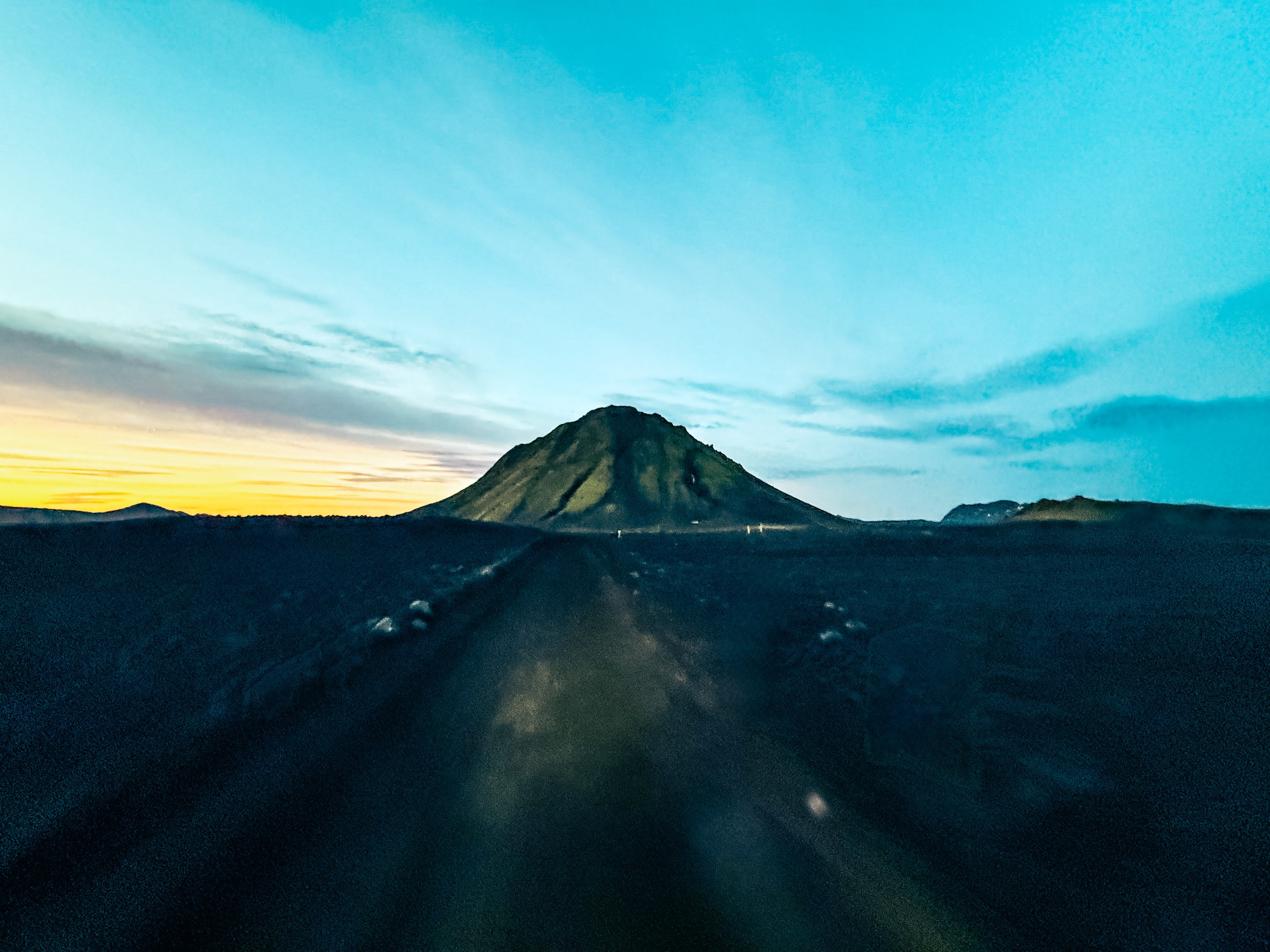 Scenic view of a volcanic mountain at sunset, with a winding road leading toward the peak under a colorful sky.