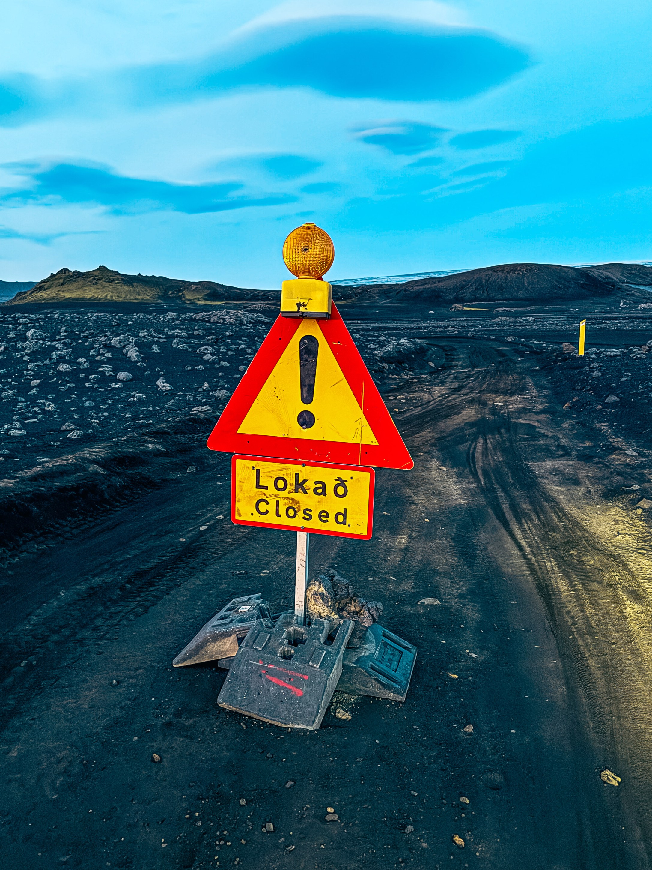 A traffic warning sign indicating 'Closed' in Icelandic, featuring a yellow background and red triangular shape, positioned on a gravel road in a volcanic landscape under a blue sky.