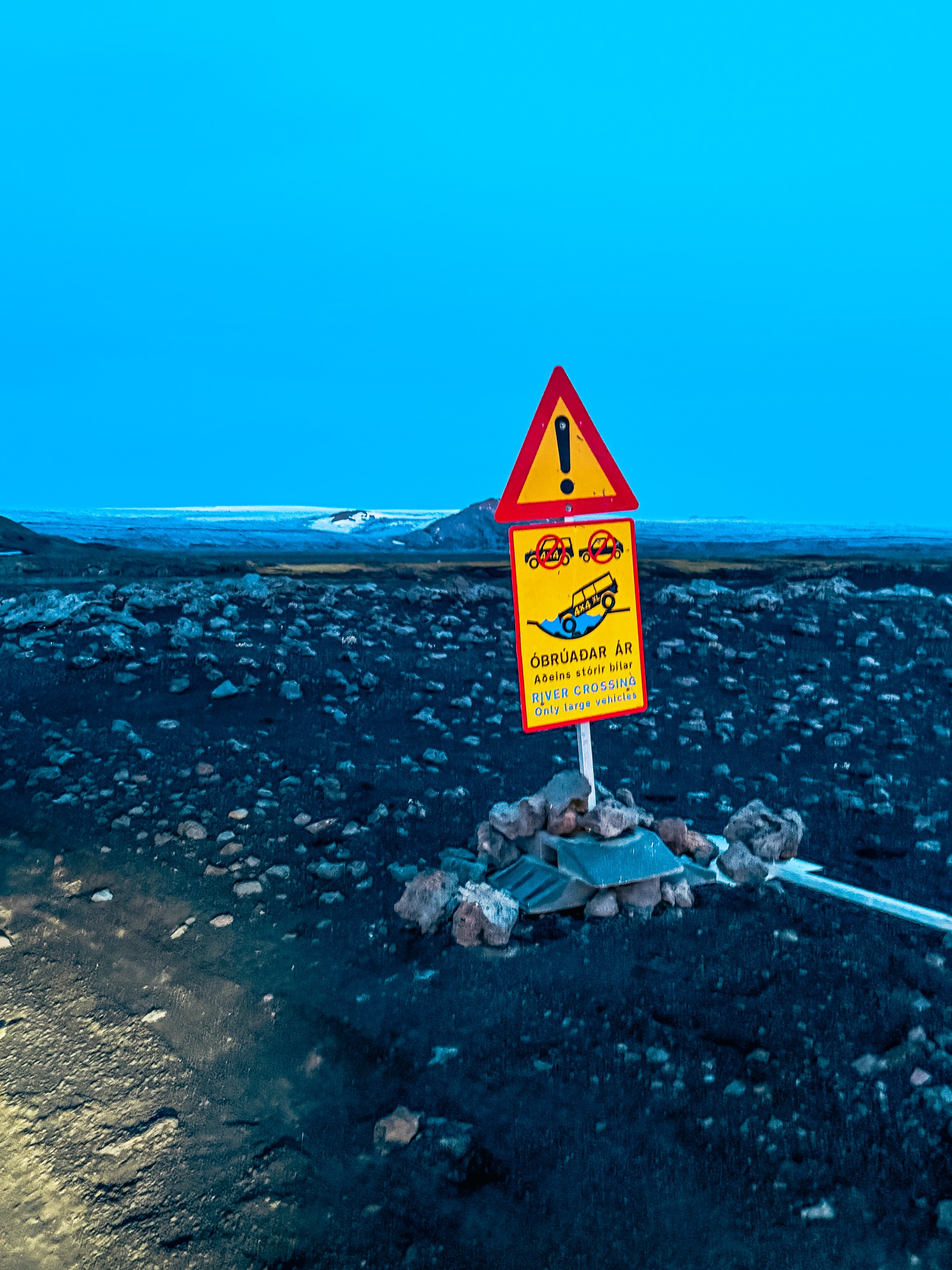 Warning sign indicating a river crossing area, advising only large vehicles can pass, set against a rocky landscape with a blue sky.
