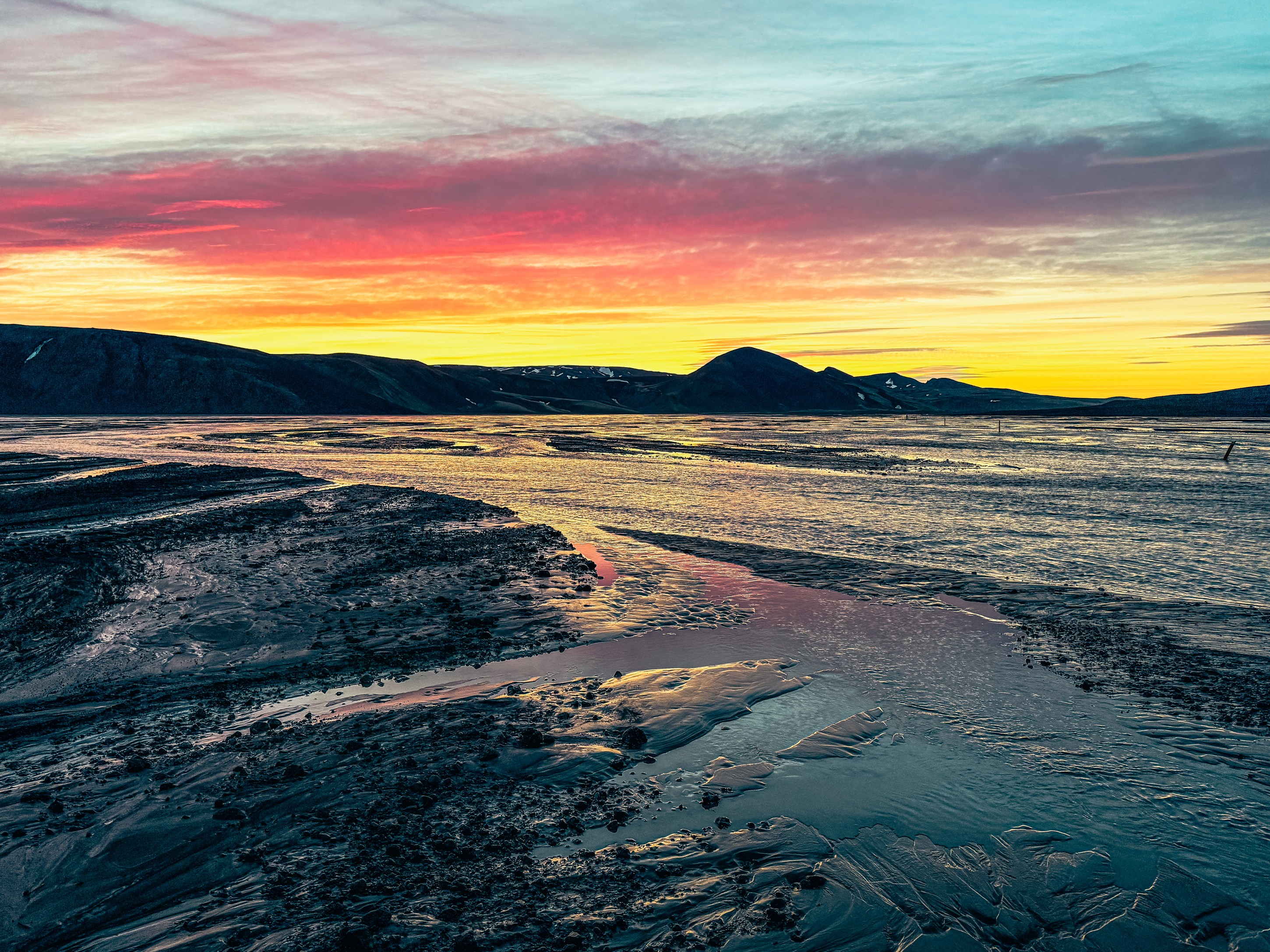 A serene landscape at sunset, featuring a winding river reflecting vibrant orange, pink, and blue hues, with dark hills in the background.