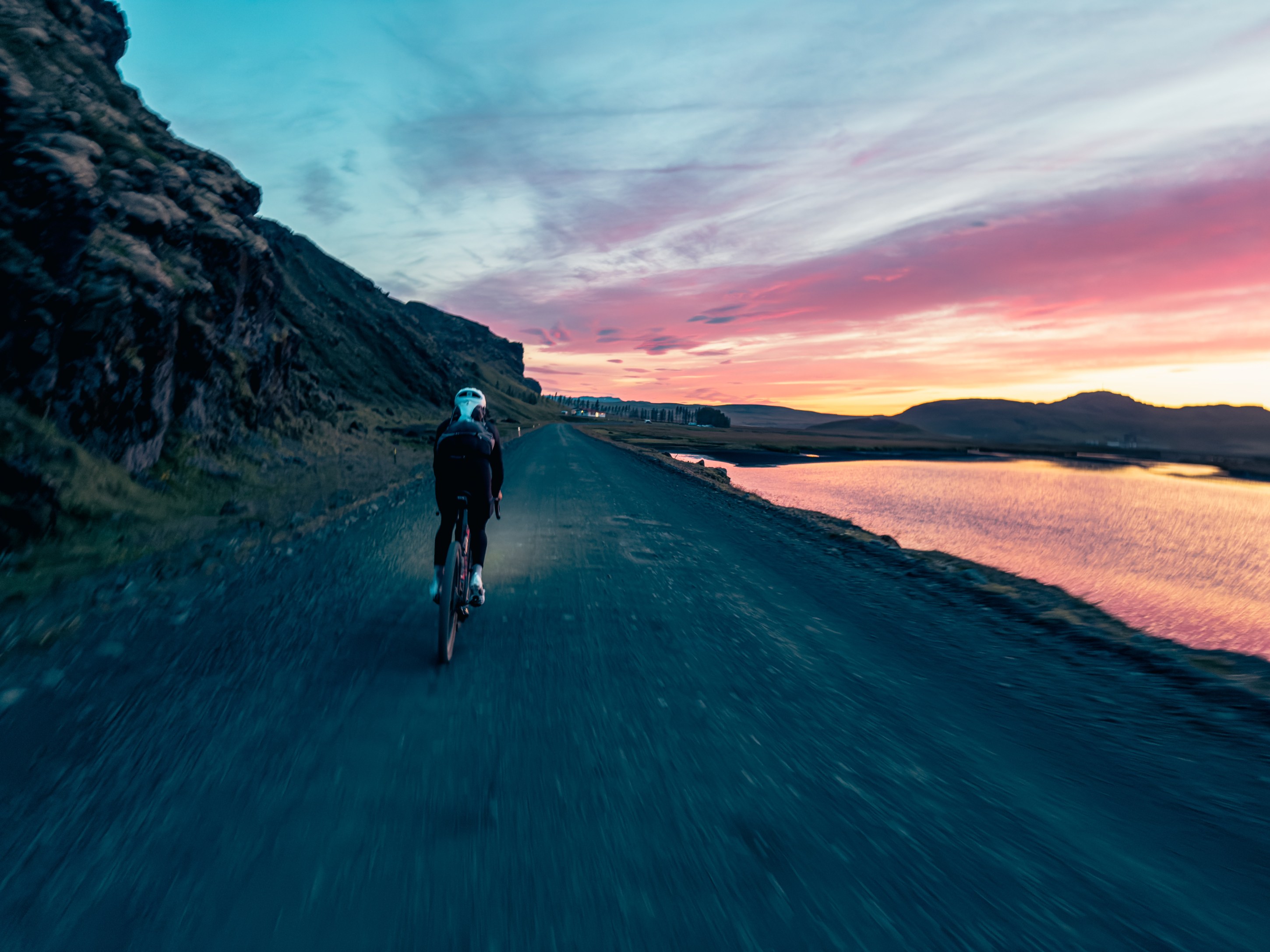 A cyclist riding on a gravel road alongside a lake during sunset, with vibrant colors in the sky and rocky cliffs in the background.