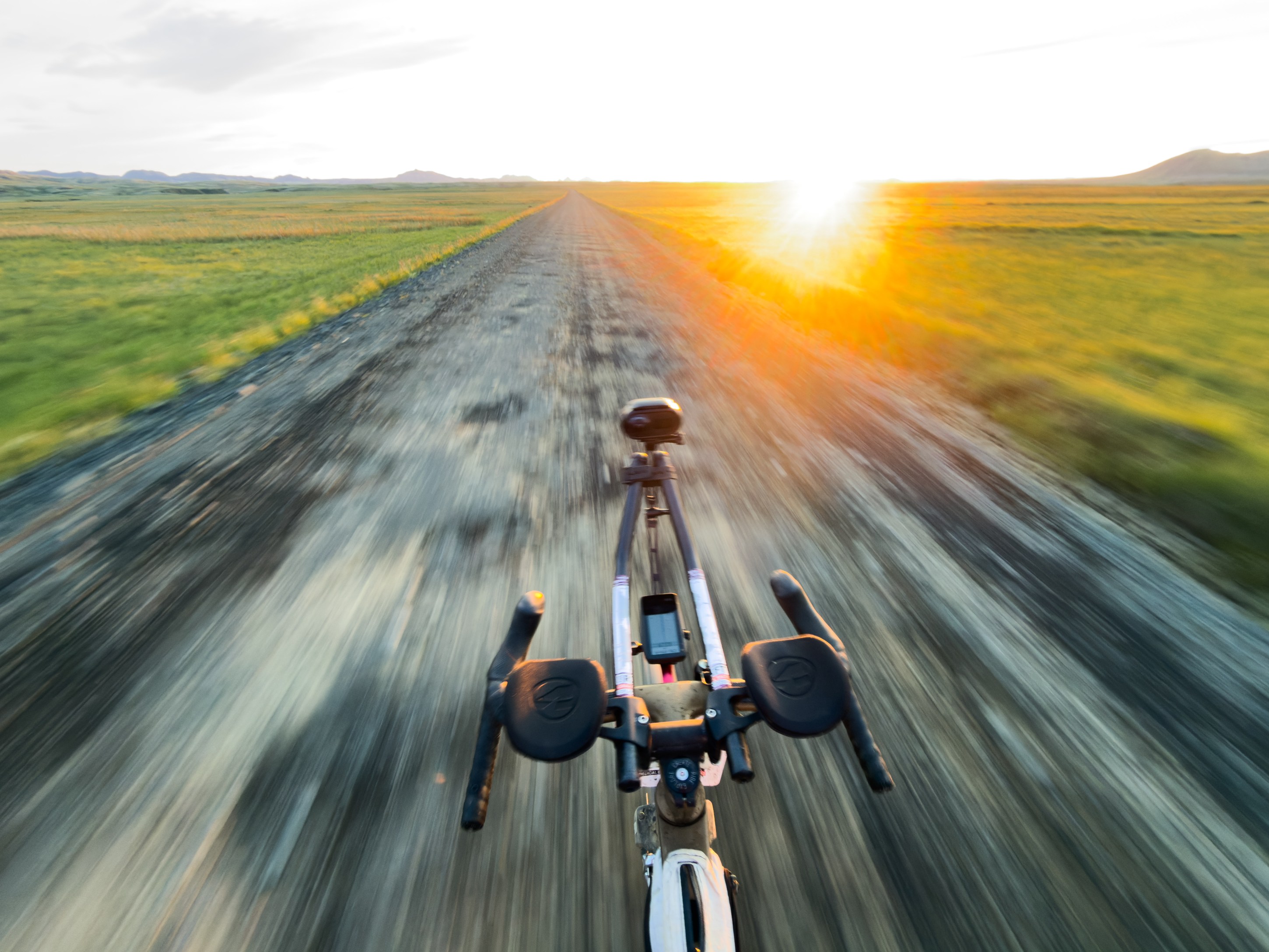 A close-up view of a bicycle's handlebars and front wheel, with a blurred dirt road extending ahead, bathed in warm sunlight during sunset.