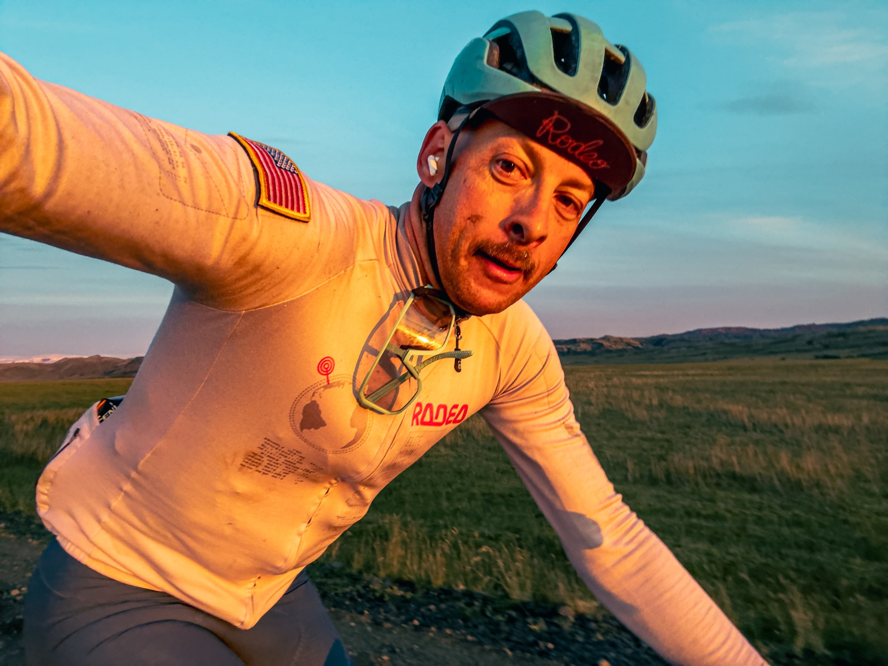 A cyclist wearing a light-colored jersey with an American flag patch and sunglasses, riding on a dirt road during sunset with grassy hills in the background.