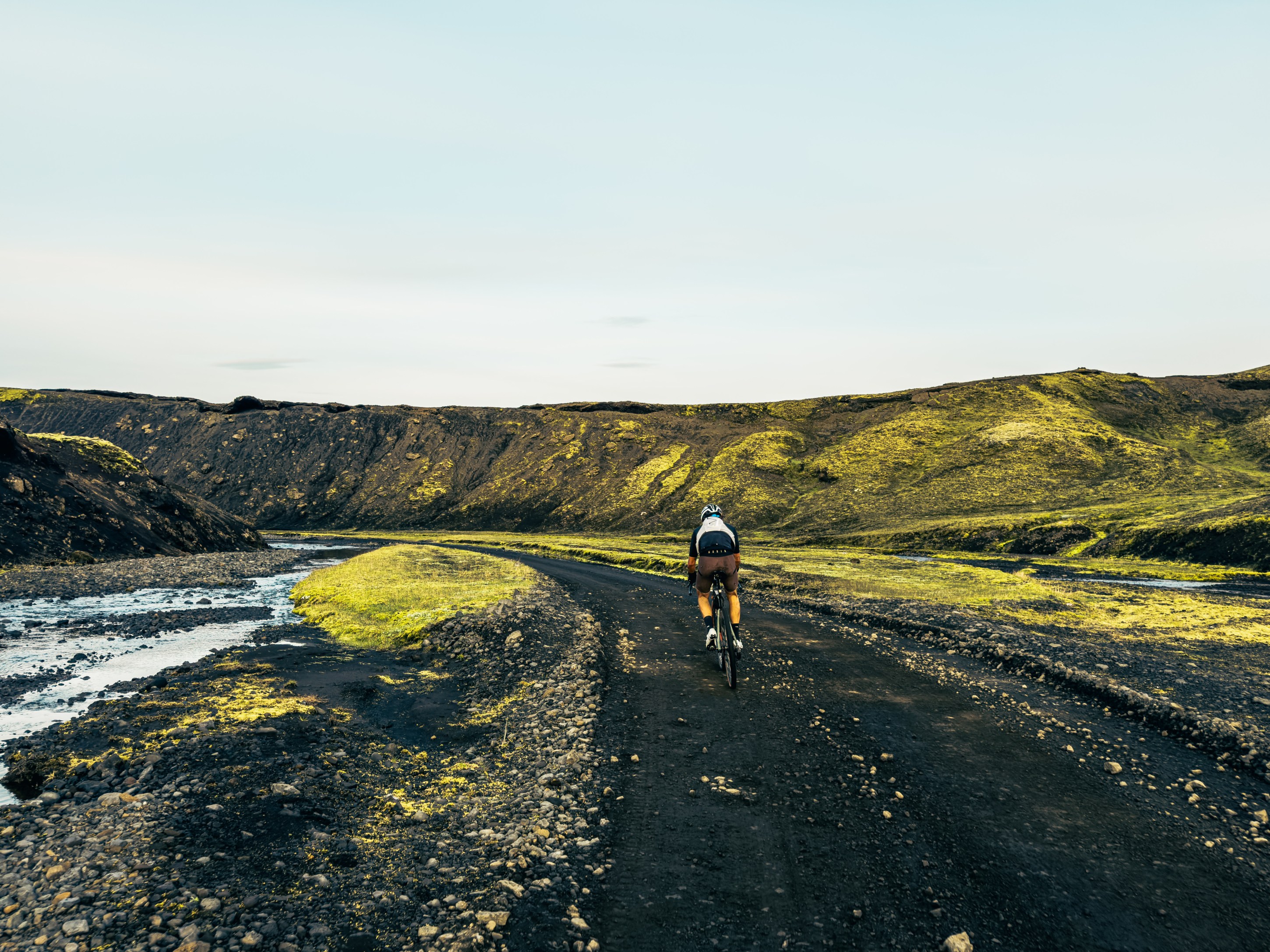 A cyclist riding along a gravel path through a rugged landscape with green hills and a winding river.