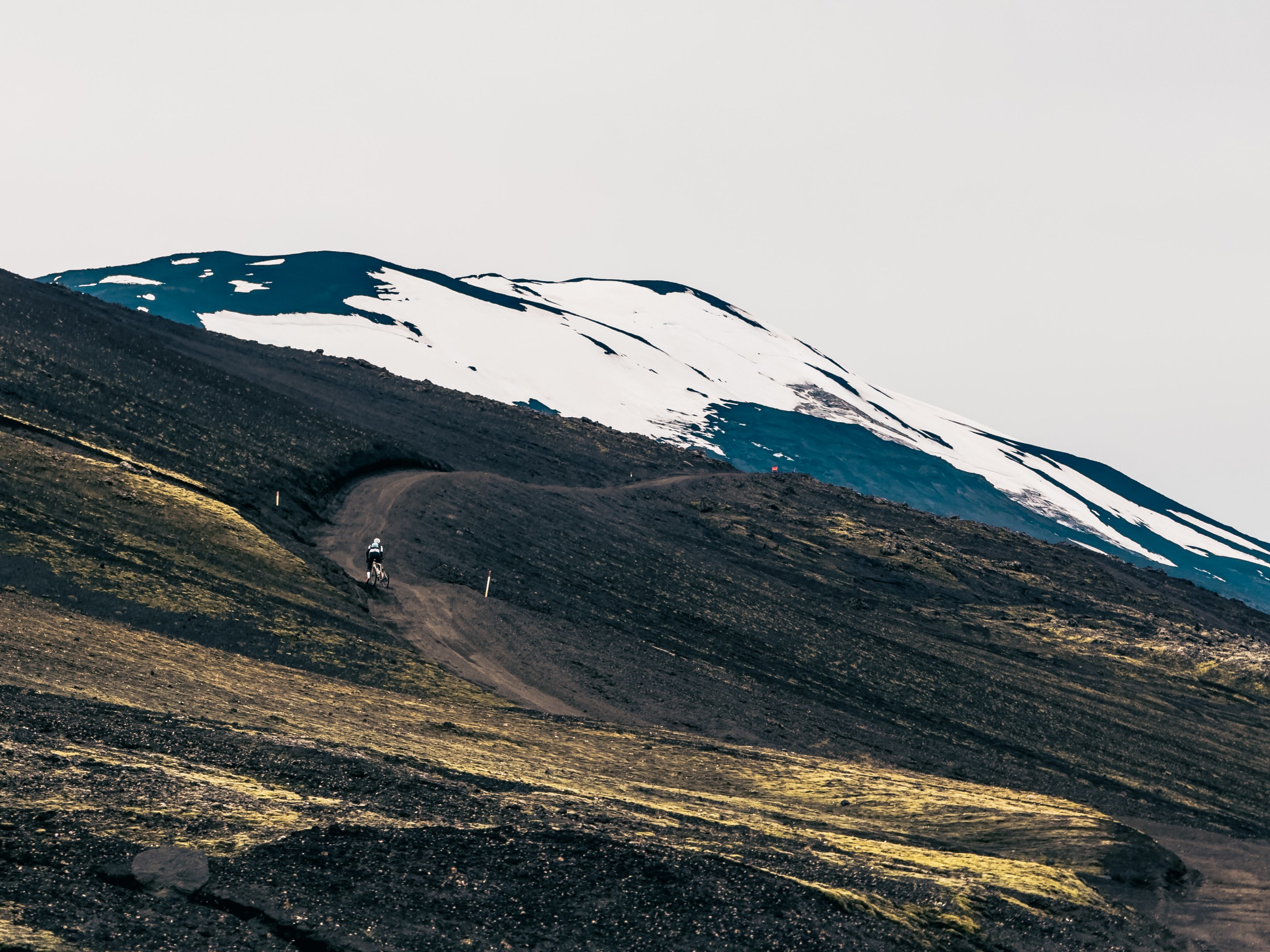 A remote mountain landscape featuring black volcanic soil and patches of snow, with a winding dirt path leading up the slope and a lone cyclist in the distance.