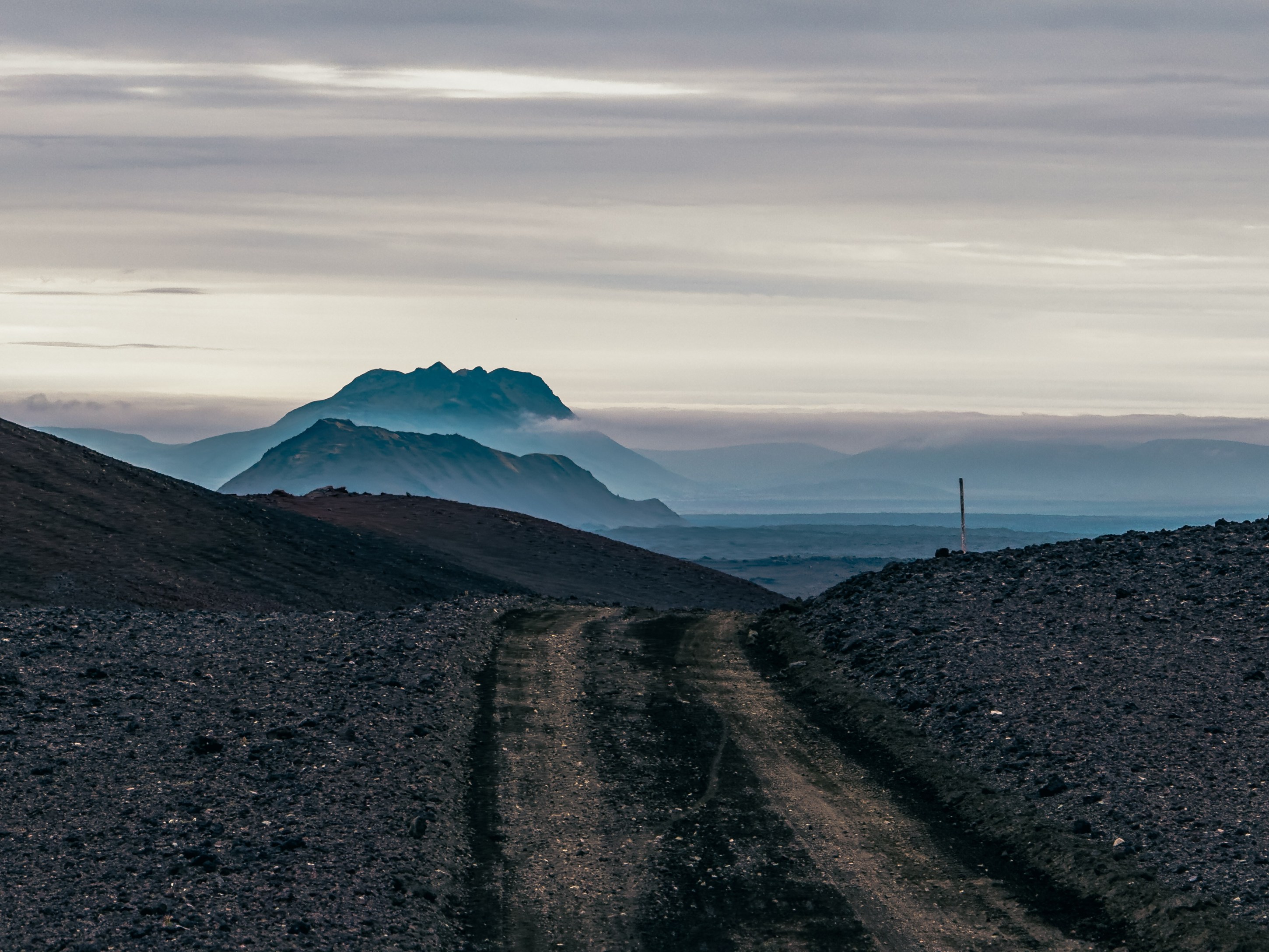 A winding dirt road leads through a rugged landscape with mountains in the background under a cloudy sky.