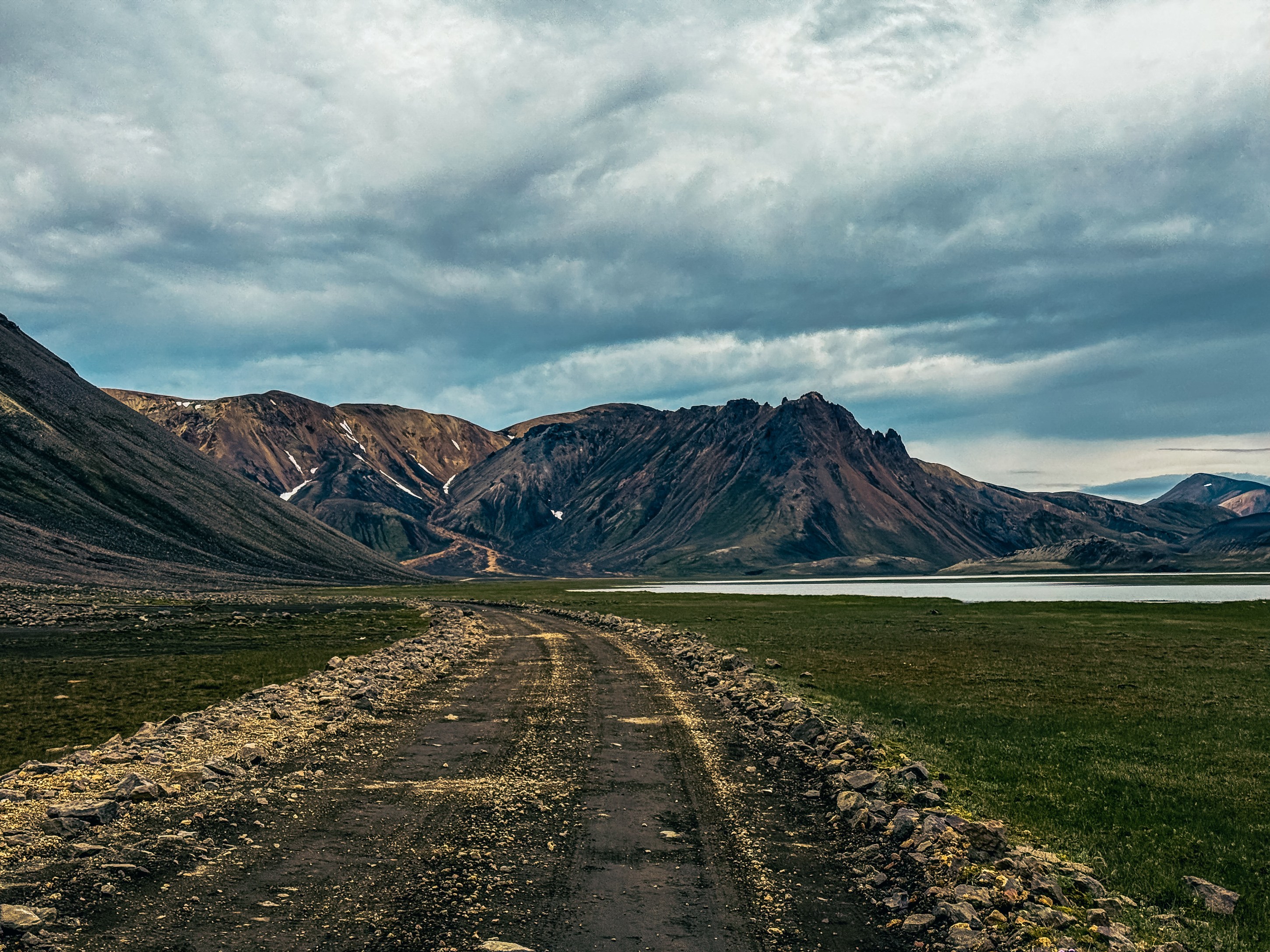 A rugged dirt road winding through a mountainous landscape with lush green grass and a calm body of water in the background, under a cloudy sky.