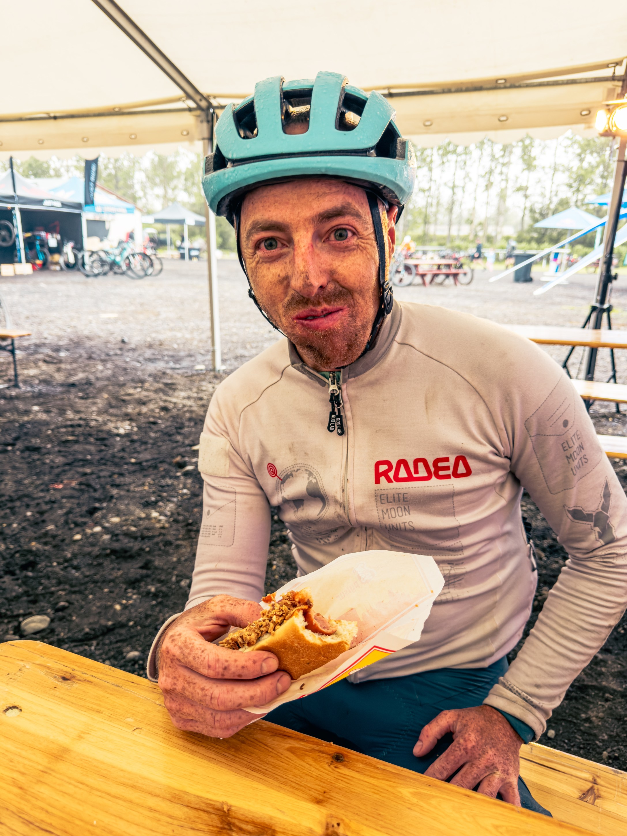 A cyclist wearing a turquoise helmet and a long-sleeve jersey, covered in dirt, sits at a wooden table holding a sandwich. The background shows a camping area with tents and bikes.
