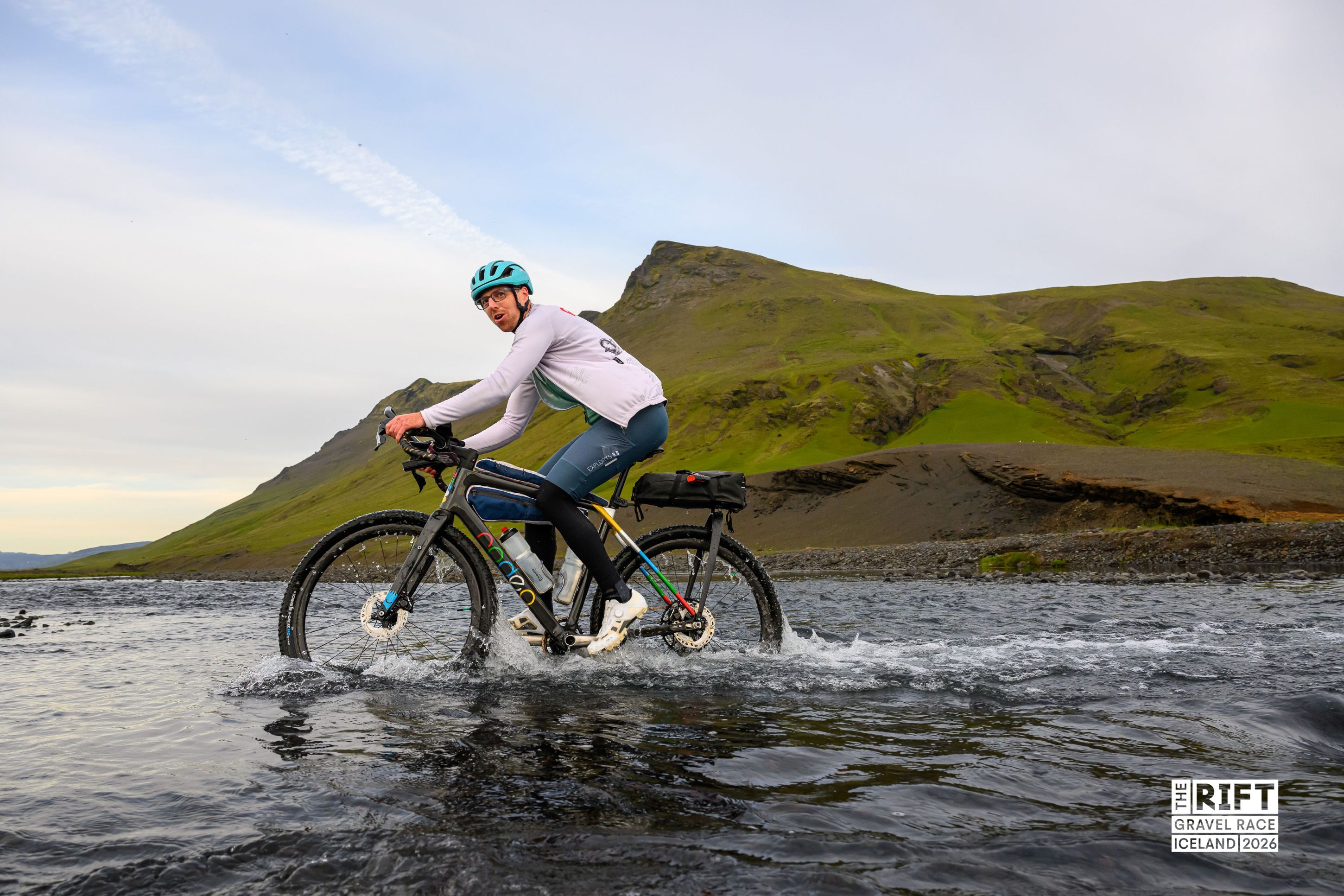 A cyclist riding through a shallow river with mountains in the background during a gravel race in Iceland.