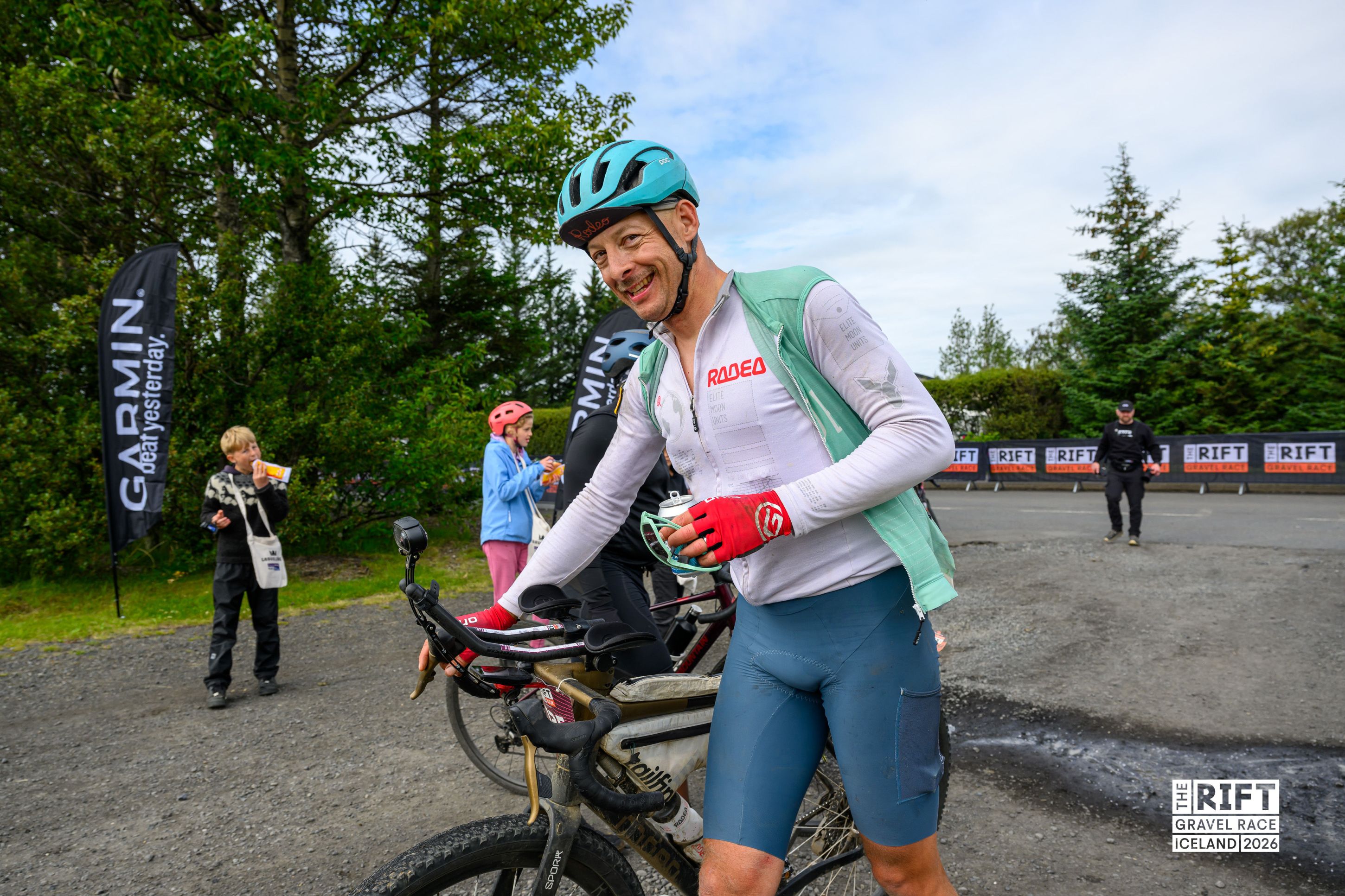 A cyclist wearing a turquoise helmet and a green vest smiles as he stands next to his bike after a gravel race in Iceland. In the background, a group of people, including children, are seen near a Garmin banner and trees.