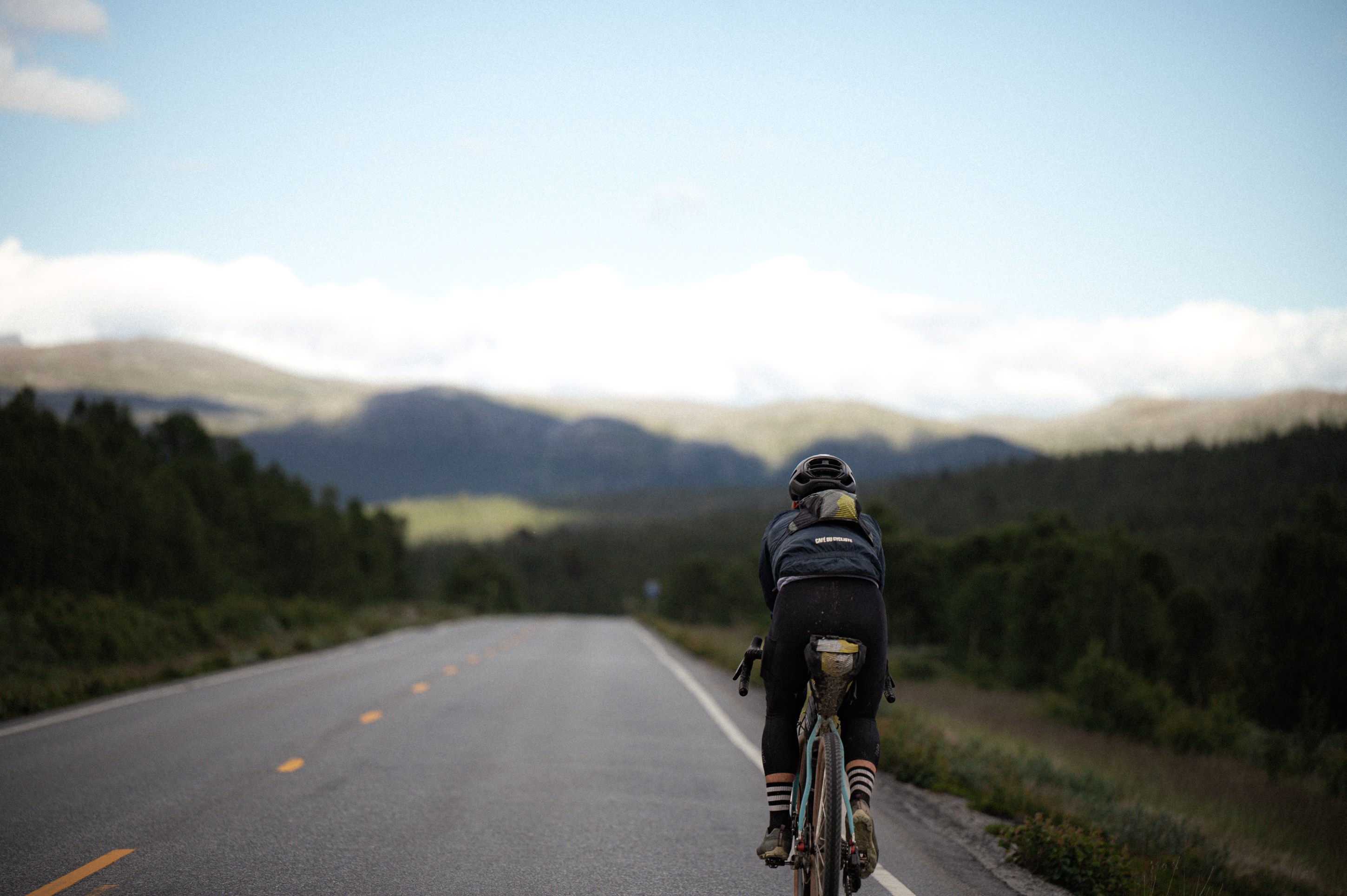 A cyclist riding on a long, empty road surrounded by mountains and trees under a partly cloudy sky.