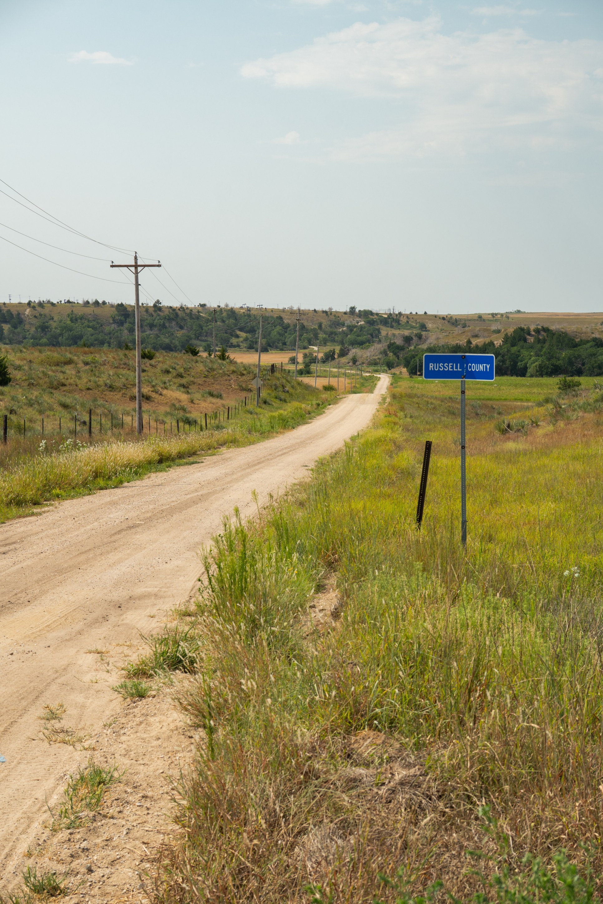 A dirt road curving through a rural landscape, with a road sign indicating 'Russell County' on the right side, surrounded by fields and greenery.