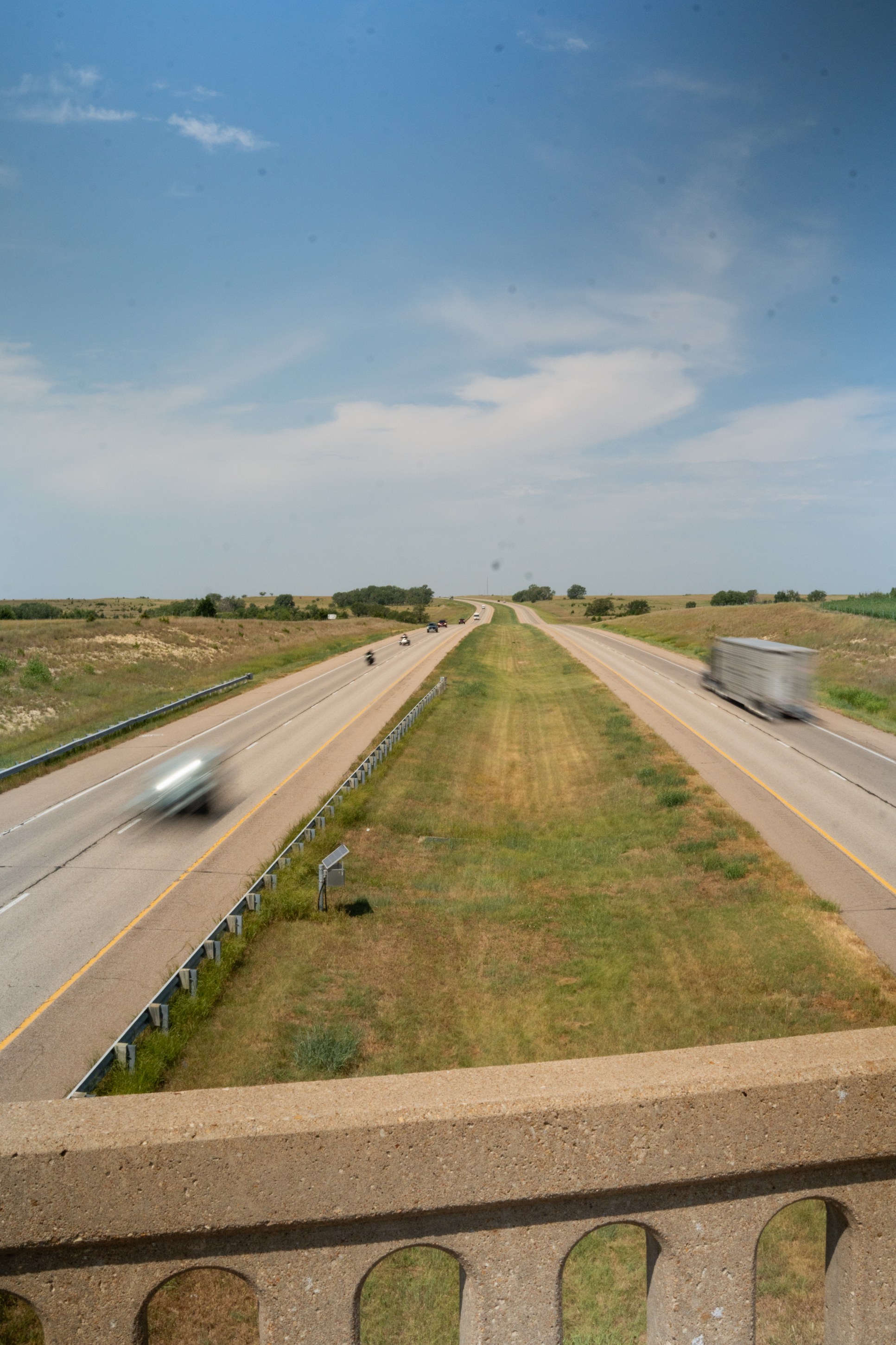 View from above a highway with light traffic, featuring vehicles traveling in both directions with a clear blue sky and green grass along the roadside.