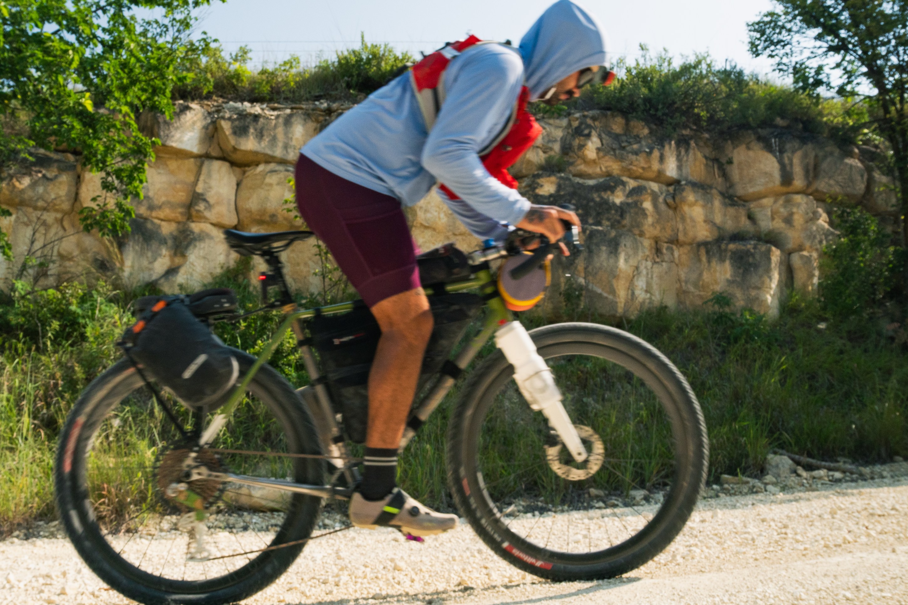 A cyclist in a blue long-sleeve shirt and red backpack rides a bike on a gravel path, with rocky terrain and greenery in the background.