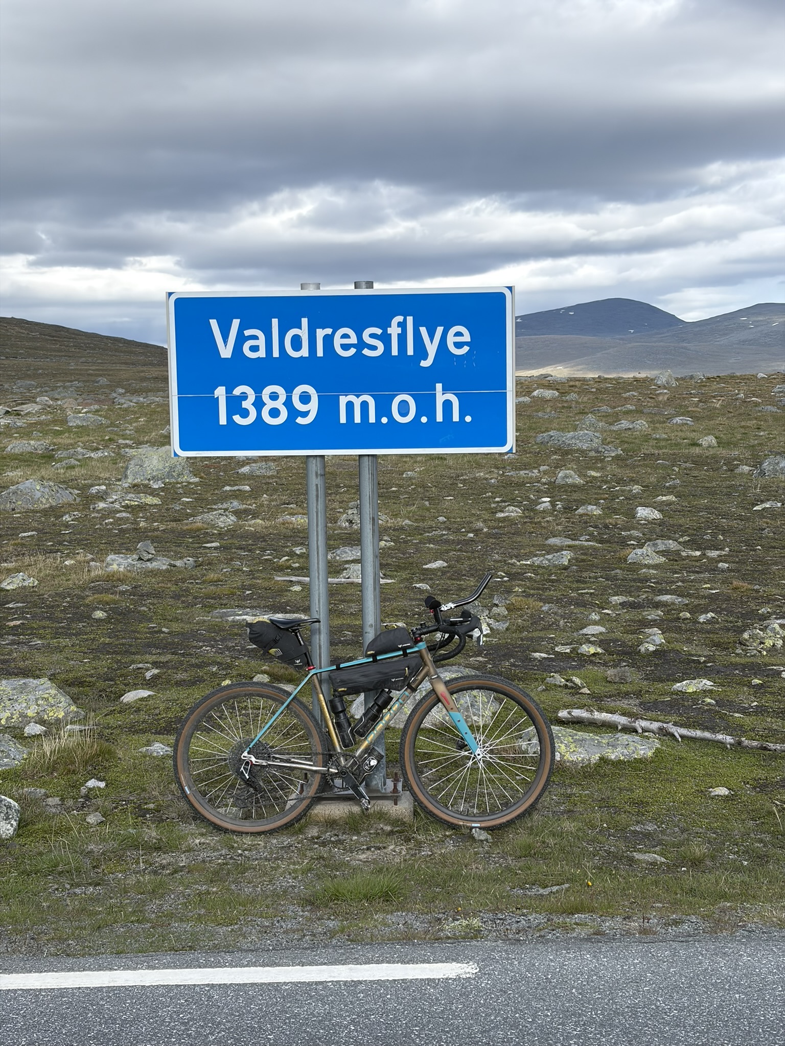 A blue road sign reading 'Valdresflye 1389 m.o.h.' stands beside a bicycle on a rocky terrain under a cloudy sky.