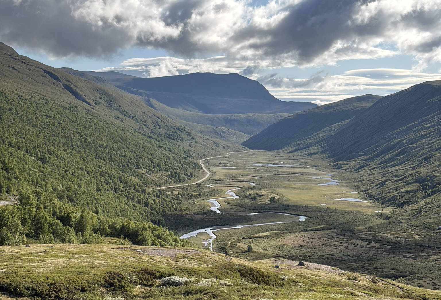 A panoramic view of a scenic valley surrounded by mountains, featuring lush greenery, winding rivers, and a clear blue sky with some clouds.