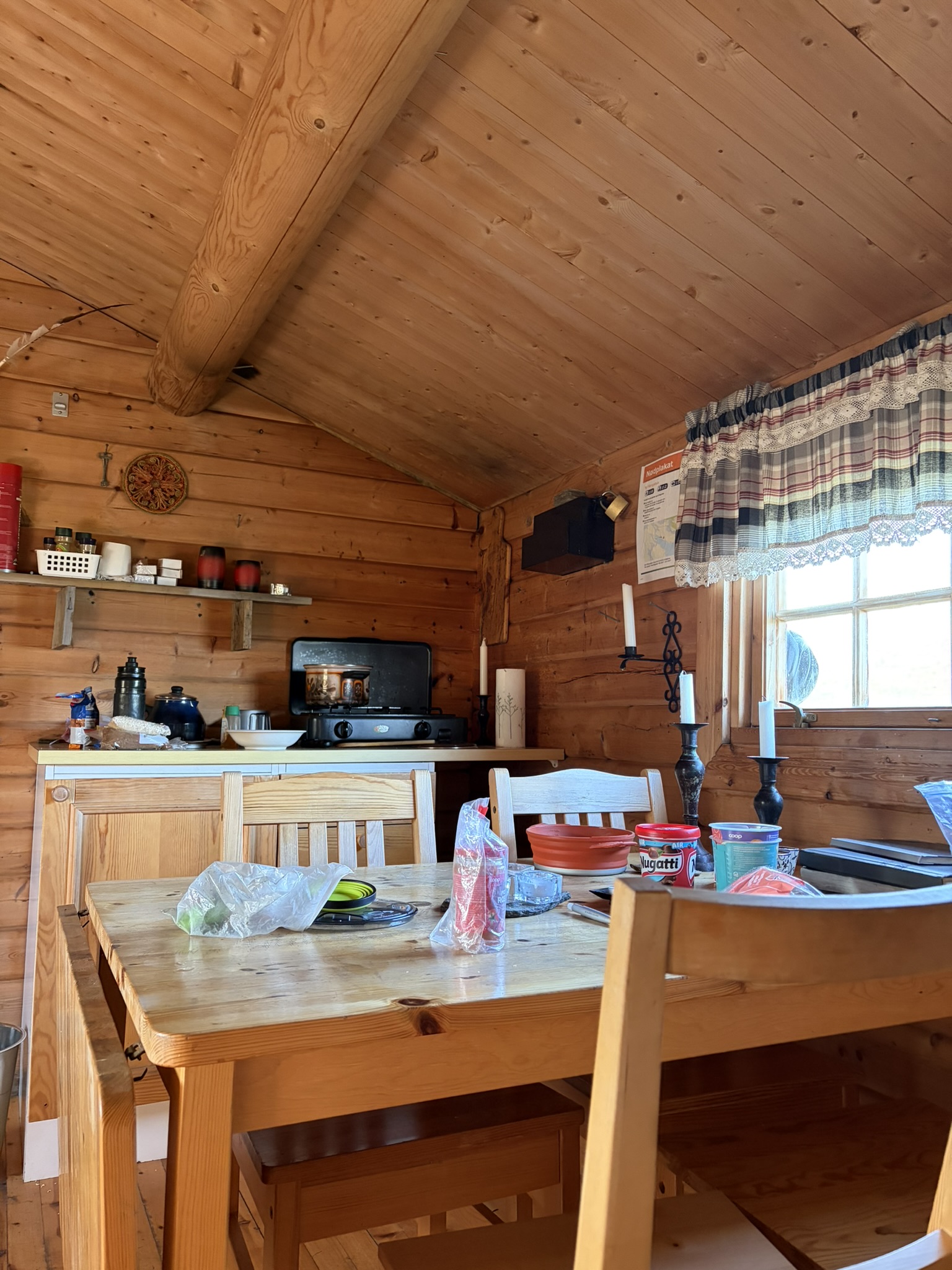 Interior of a rustic wooden cabin kitchen, featuring a wooden dining table with various items on it, including plastic bags and containers, a stove in the background, and wooden beams on the ceiling.