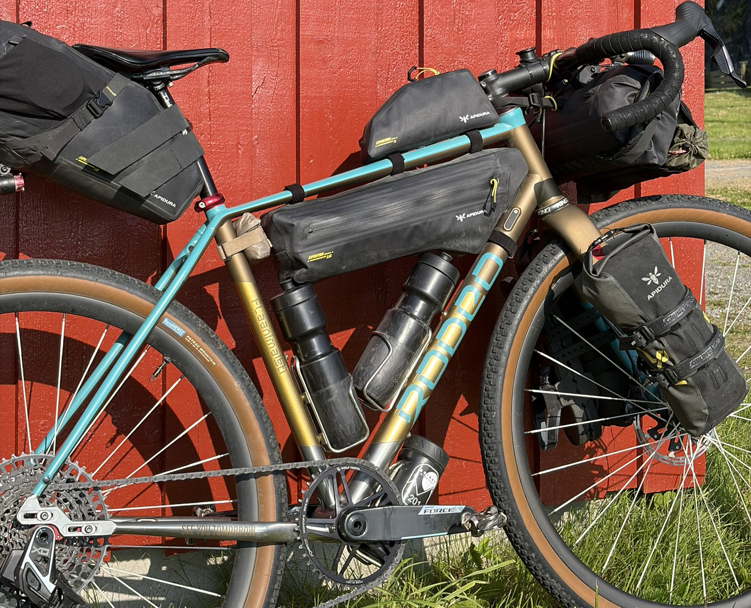 A close-up view of a bicycle equipped with multiple black bags for storage, positioned next to a red wooden wall with grass at the bottom.