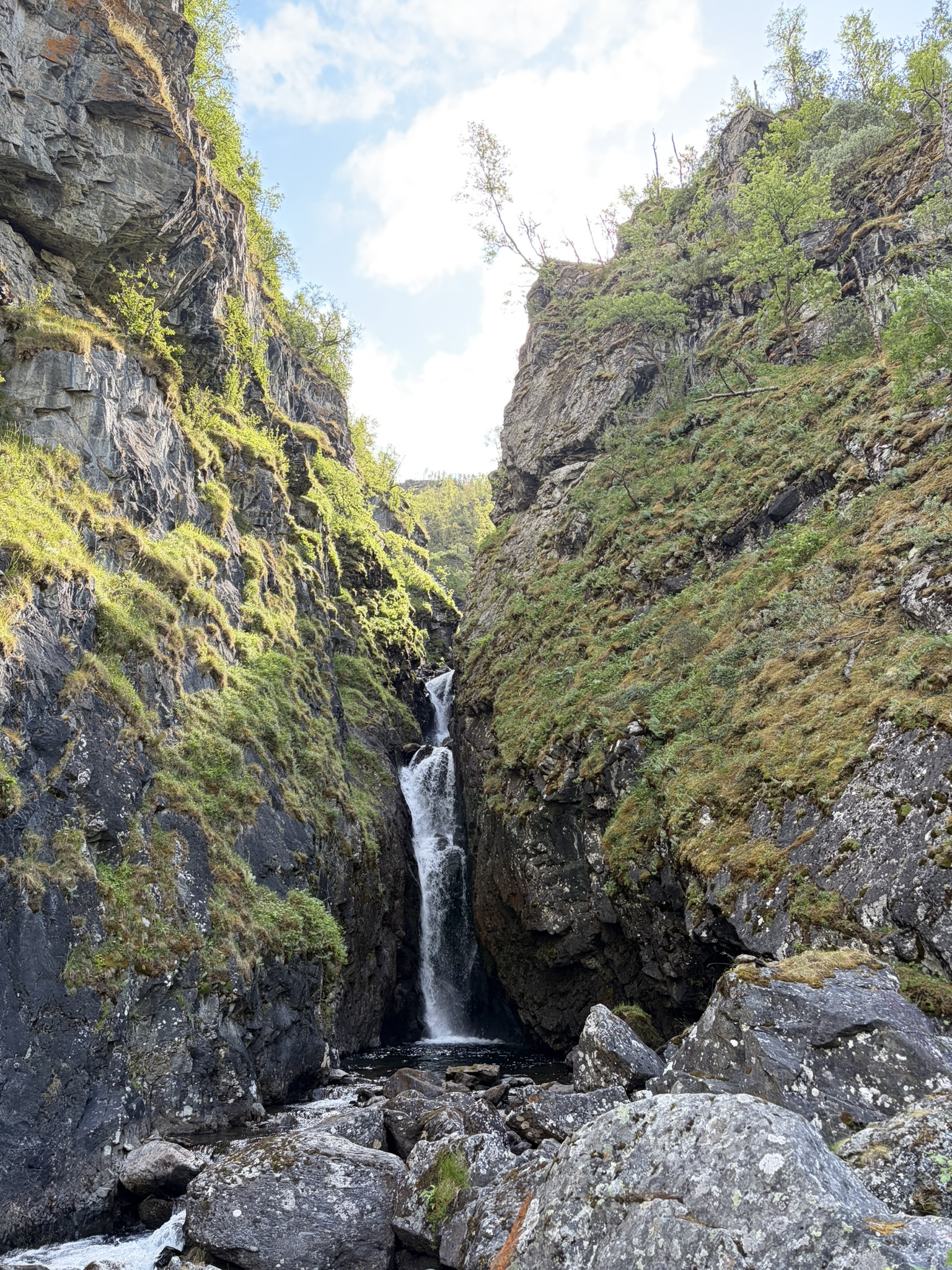 A narrow canyon with steep rocky walls, featuring a waterfall cascading down into a shallow pool surrounded by large stones and lush greenery.