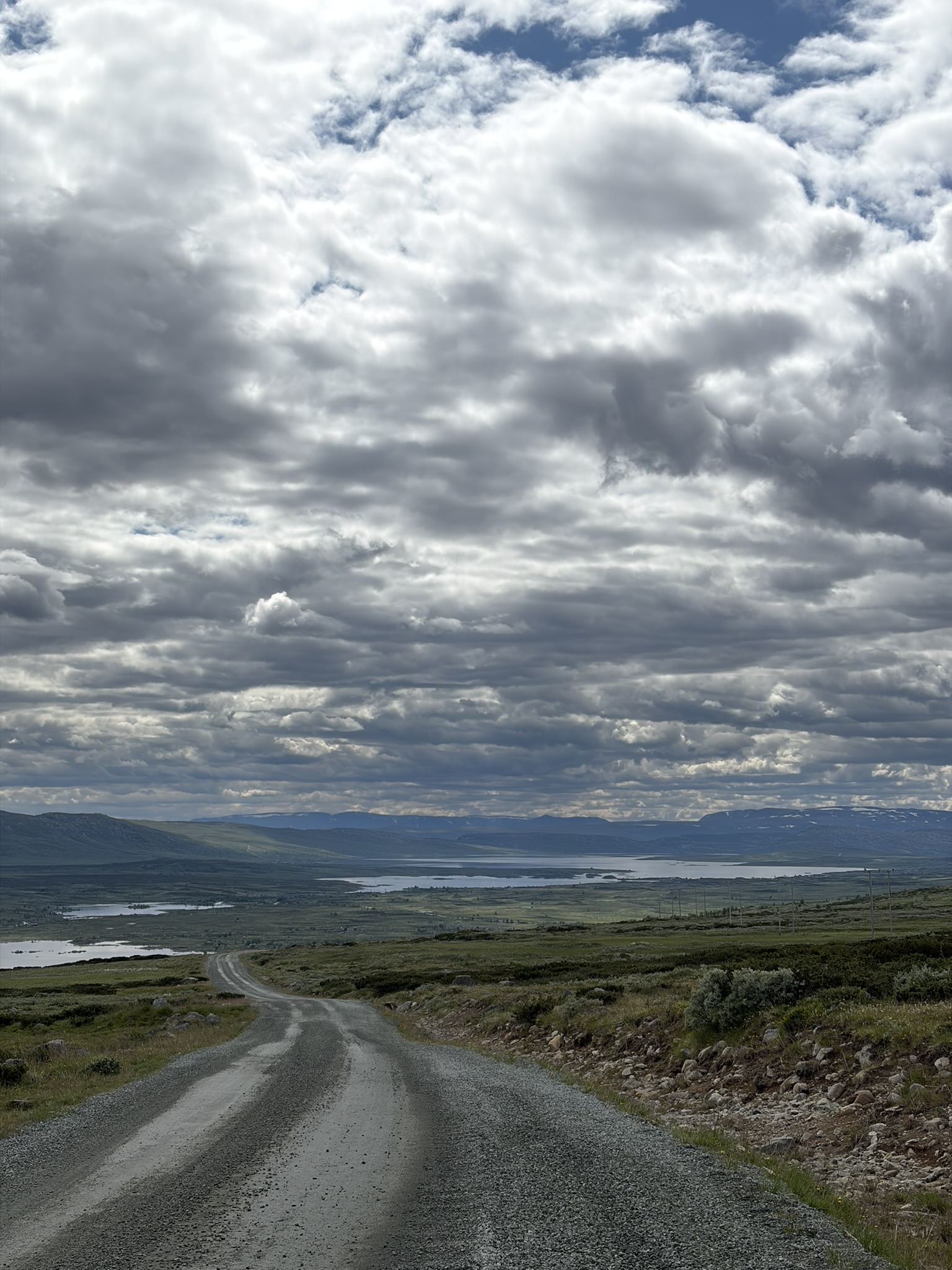 Gravel road winding through green landscape under a cloudy sky with distant hills and a lake.