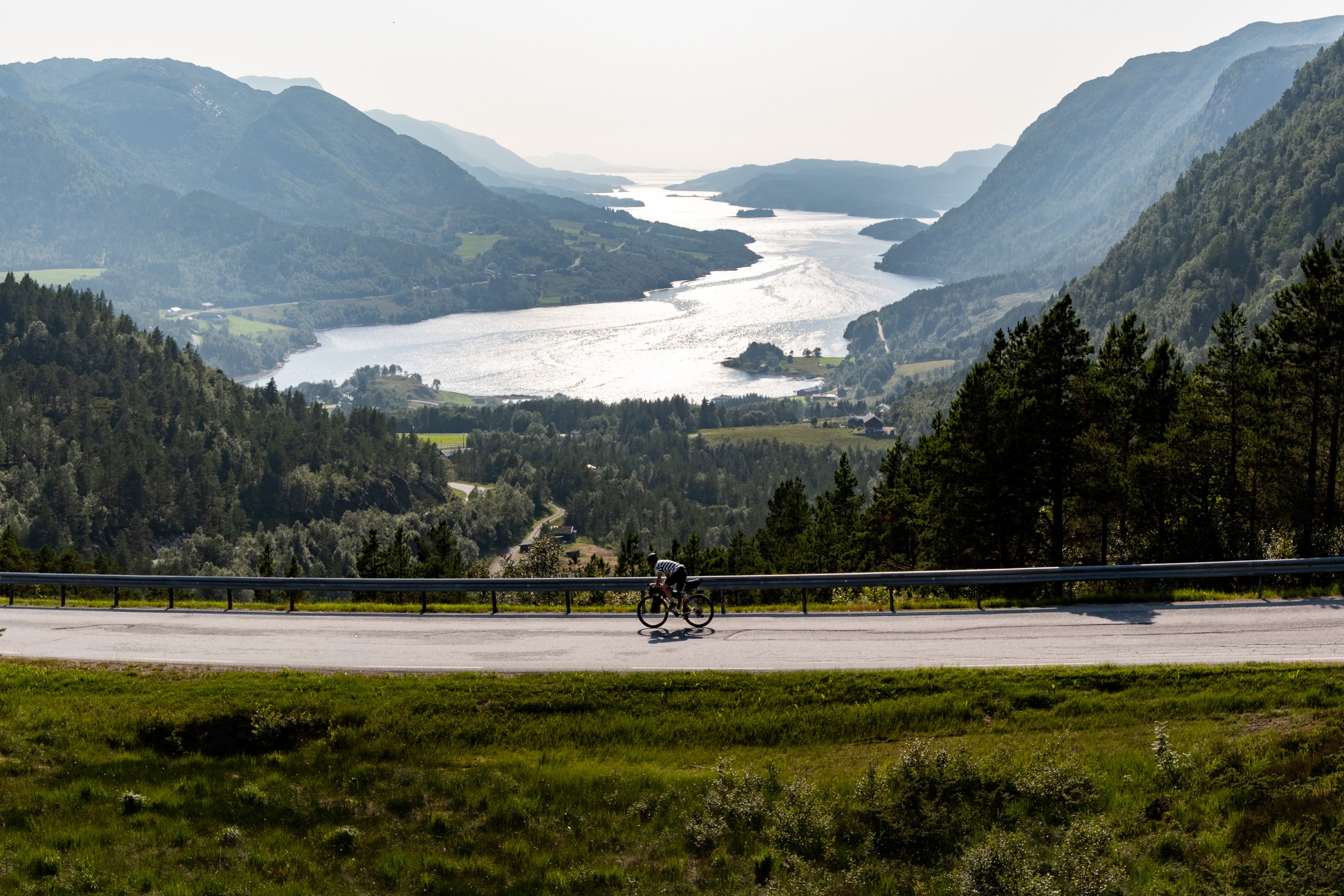 A cyclist riding on a winding road overlooking a picturesque valley with mountains and a shimmering lake in the background.