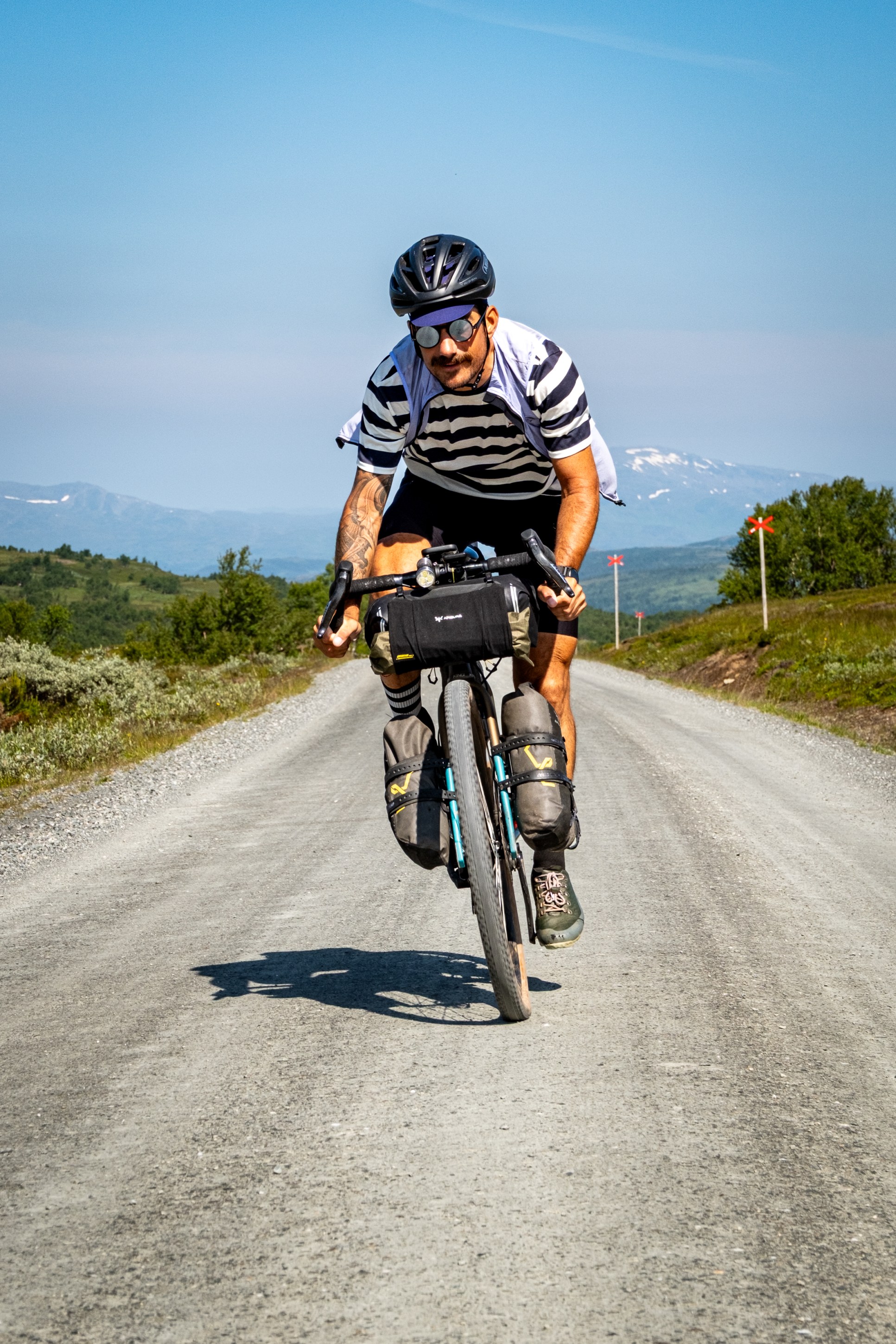 A cyclist with sunglasses and a striped shirt rides a gravel road surrounded by green landscapes on a sunny day.