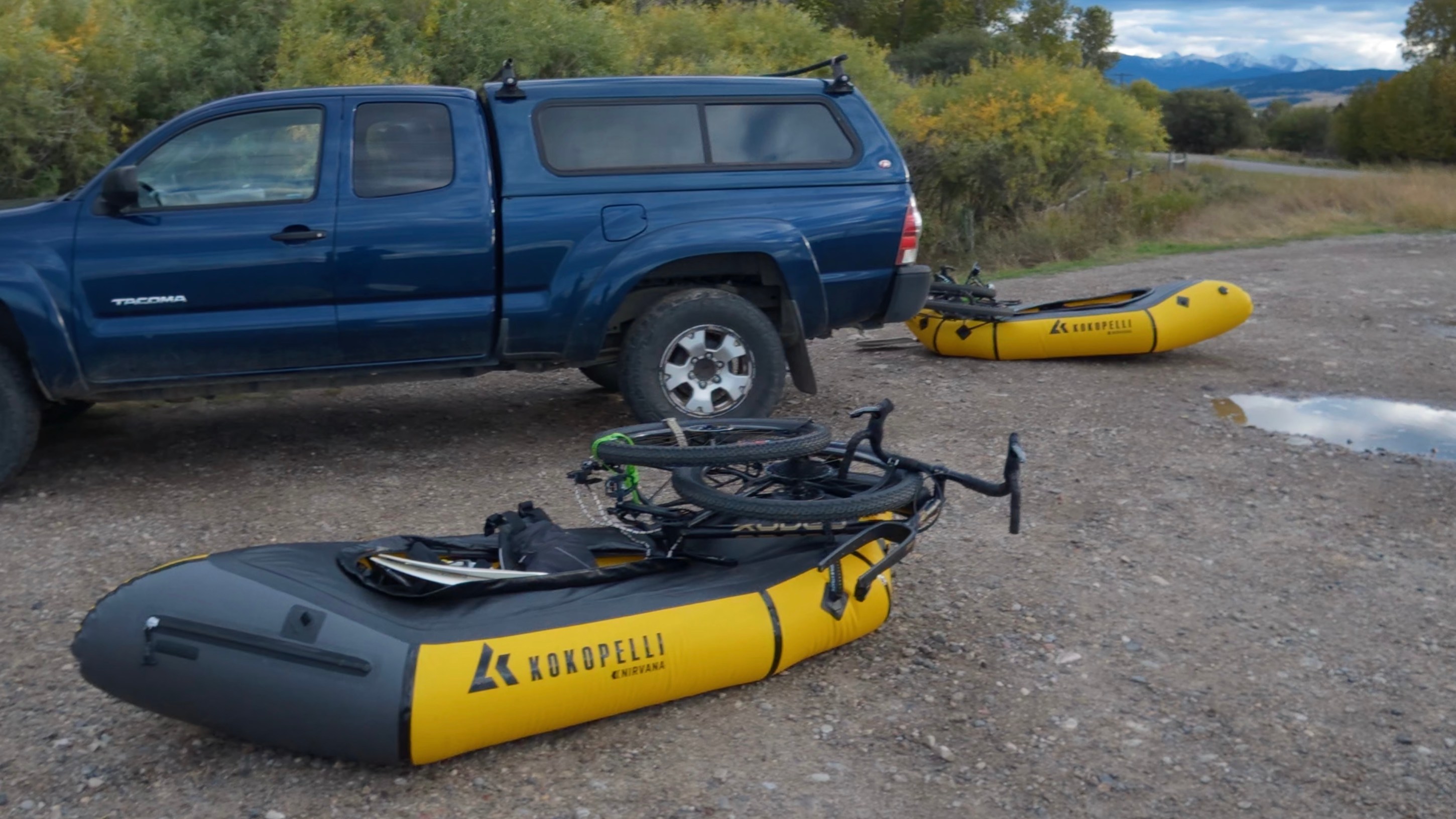 A blue pickup truck parked next to two yellow inflatable kayaks on a gravel surface, with bike frames resting on one of the kayaks and a mountainous landscape in the background.